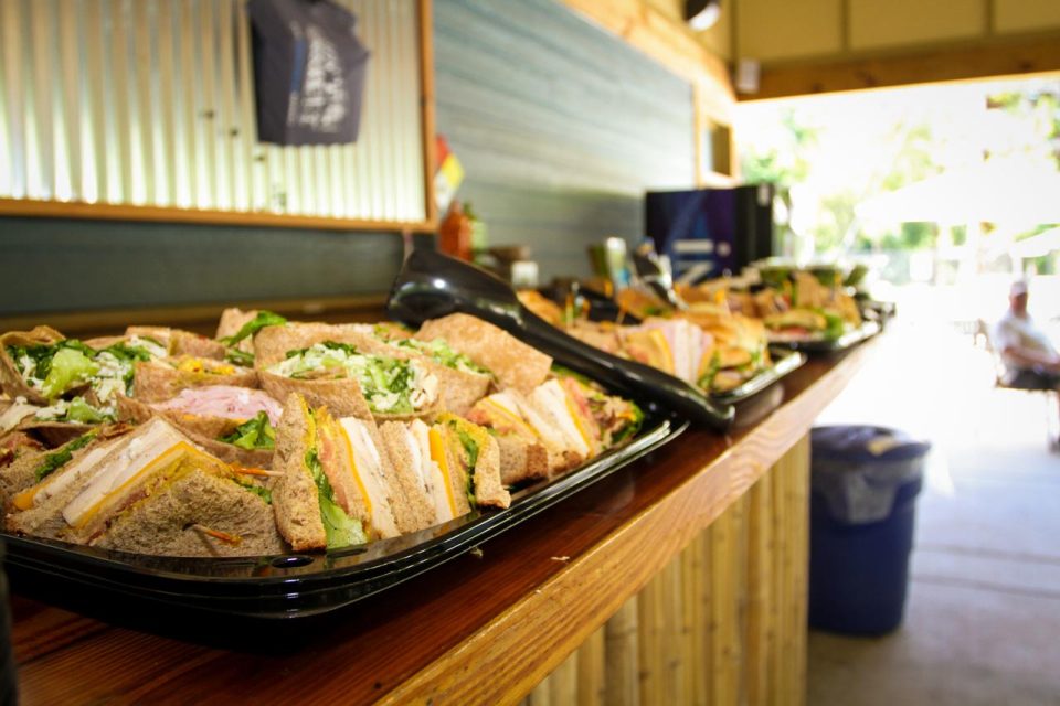 A long wooden tiki bar is lined with trays of assorted sandwiches filled with a variety of meats, cheese, and lettuce. A pair of black tongs rests on each tray.