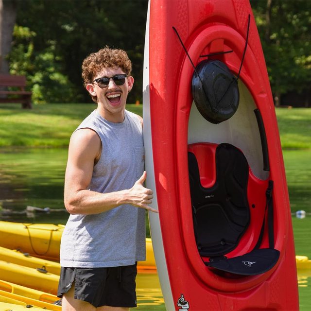 Person wearing sunglasses and a sleeveless gray shirt smiles while standing next to a red kayak held upright, with a row of yellow kayaks on the green lake at Camp Canaan in the background, and a bench and trees in the distance.