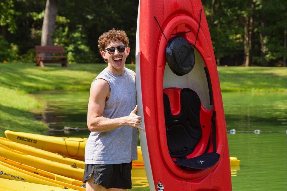 Person wearing sunglasses and a sleeveless gray shirt smiles while standing next to a red kayak held upright, with a row of yellow kayaks on the green lake at Camp Canaan in the background, and a bench and trees in the distance.