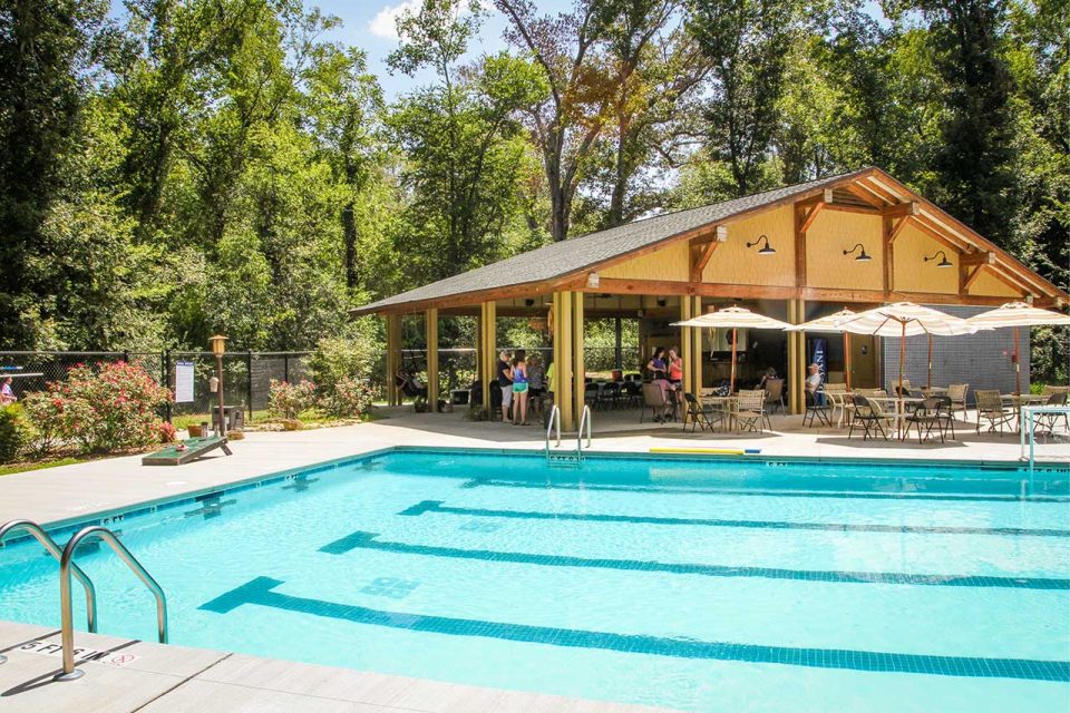 An outdoor swimming pool surrounded by a wooded area is central to Camp Canaan. Nearby, a pavilion with a sloped roof provides shade for people sitting at tables and chairs. The pool area, adorned with lounge chairs, umbrellas, and landscaped greenery, enhances the relaxing atmosphere of this summer camp and retreat center.