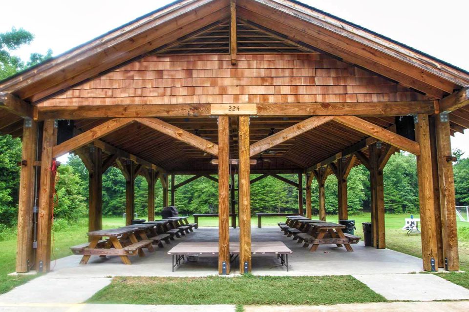 A large wooden picnic pavilion with a pitched roof and open sides, housing several picnic tables. The pavilion is situated in a green park area with trees visible in the background.