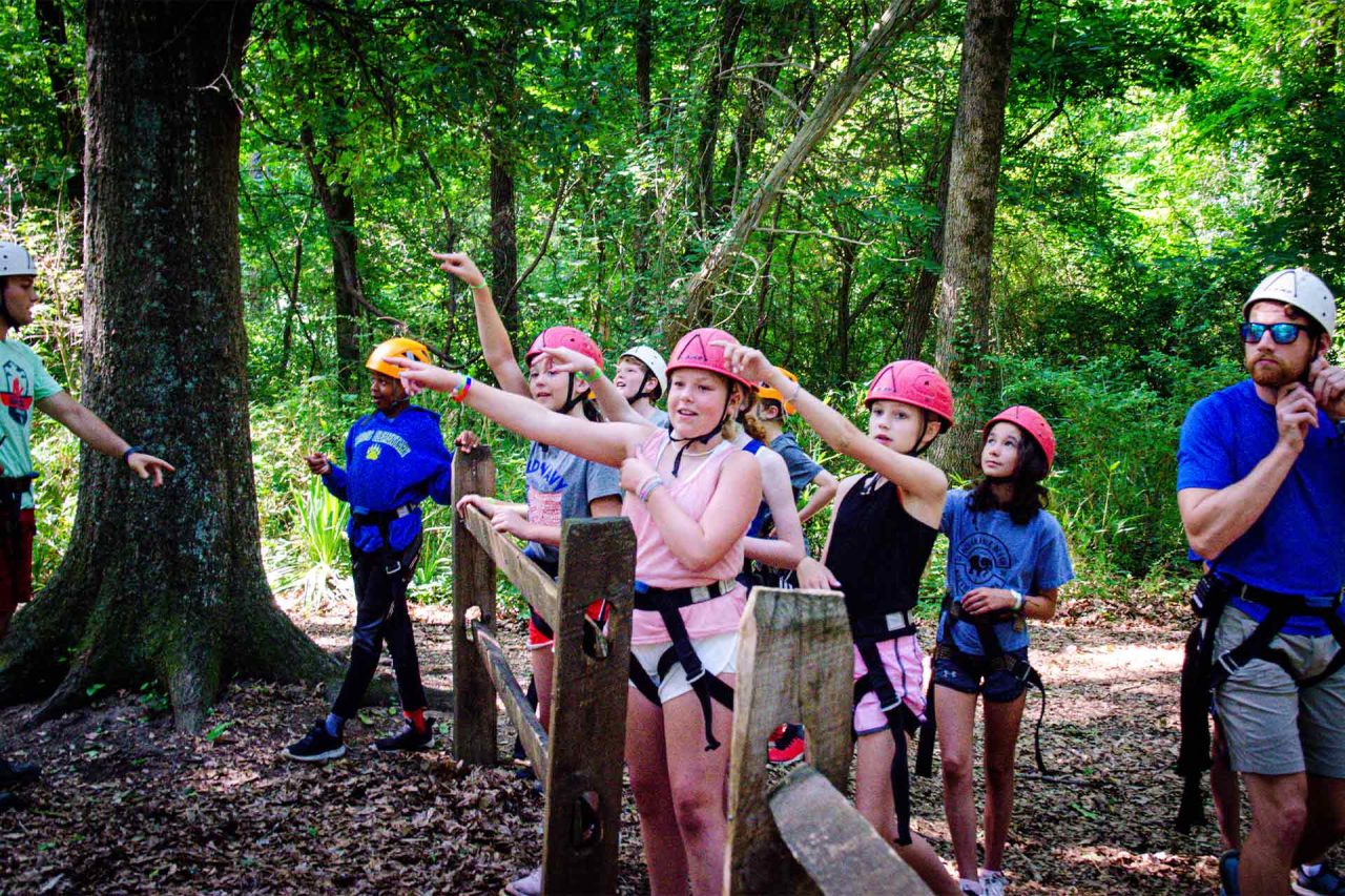 A group of children, wearing helmets and harnesses, stand in a wooded area by a fence at the summer camp. They are looking and pointing towards something, possibly getting instructions for the zipline canopy tour. An adult wearing a helmet and harness stands nearby.