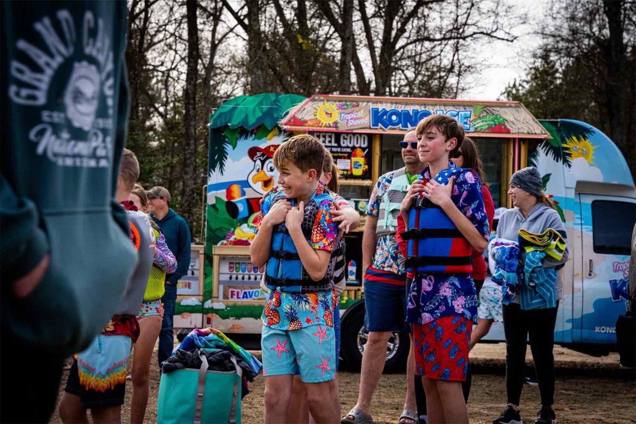 A group of children and adults stand in front of a colorful Kona Ice truck in front of Lake Canaan. The children are wearing life jackets and vibrant summer clothes, while an adult in a hoodie stands nearby. Trees and other camp-like surroundings can be seen in the background.