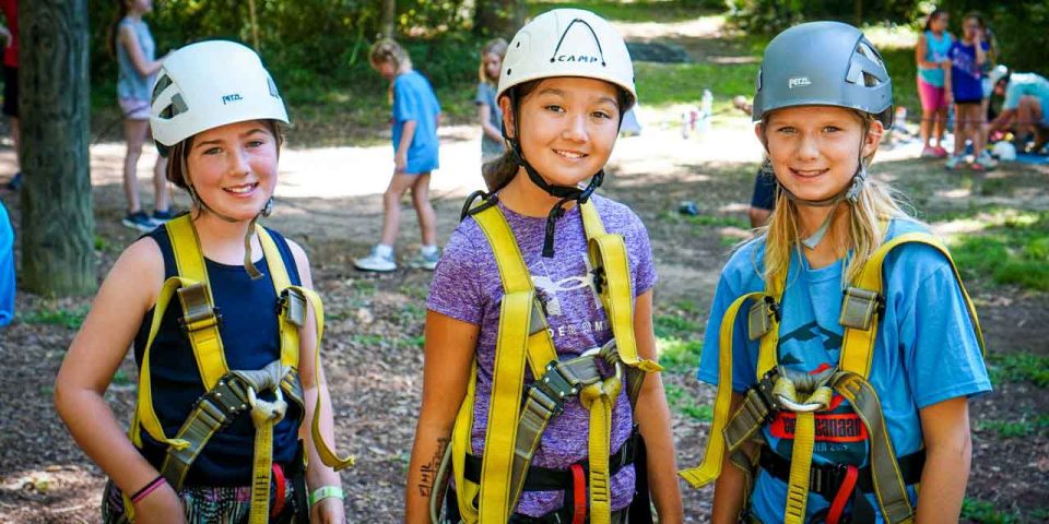 three girl campers in helmets and harnesses get ready to tackle the high ropes course at Camp Canaan