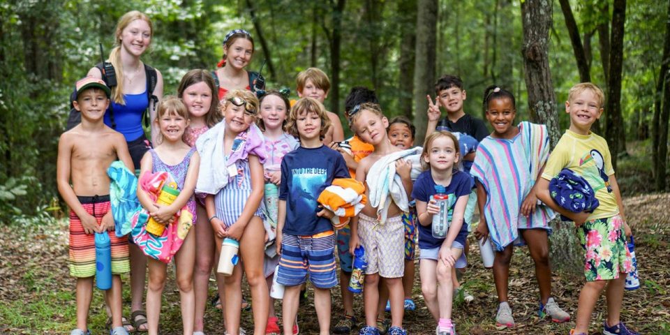 A group of smiling campers and two adults stand together outdoors in a wooded area wearing swimsuits and holding towels. The background is lush with trees and foliage, suggesting a fun day of summer camp.