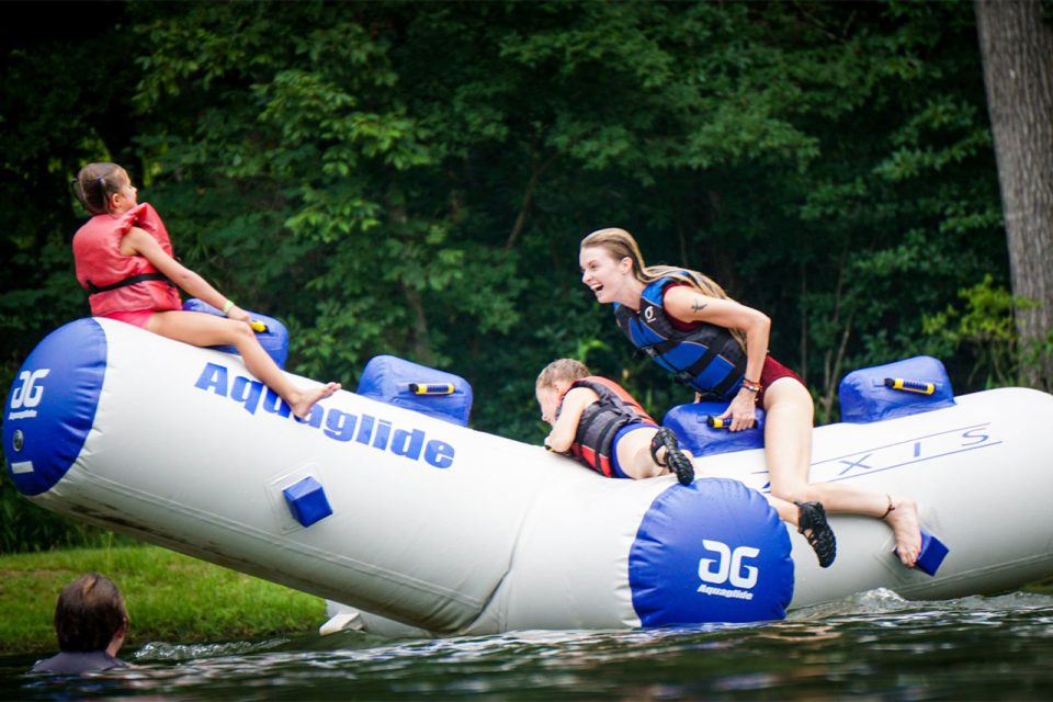 Three people, wearing life vests, are playing on an inflatable white Aquaglide water toy in the lake during a summer camp.