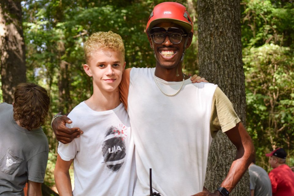 Two people stand outdoors in a wooded area getting ready for an outdoor adventure activity during summer camp. One, with short curly hair, wears a white t-shirt and smiles slightly. The other, wearing glasses, a red helmet smiles broadly with an arm around the other's shoulders. Trees fill the background.