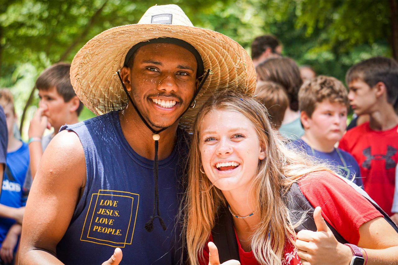 Two smiling young adults giving thumbs up. The man on the left is wearing a wide-brimmed straw hat and a sleeveless shirt that says "Love Jesus. Love People." The woman on the right has long blonde hair and is wearing a red shirt. The lively atmosphere resembles a summer camp, with people in the background.