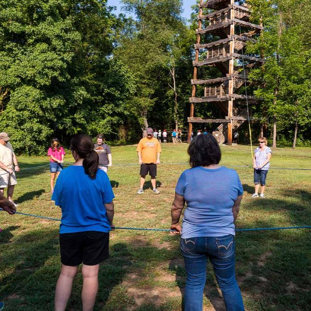 A group of people stand in a circle outdoors at Camp Canaan, holding sections of a blue rope. They are in a grassy area surrounded by trees, with the Zipline Canopy Tour tower in the background.