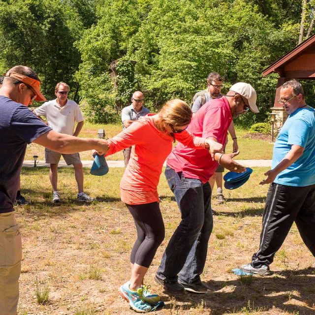 A group of people at Camp Canaan are participating in an outdoor team-building activity. Trees and a building are in the background.