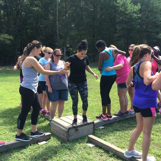 A group of women in athletic wear engage in an outdoor team-building exercise on the sports fields at Camp Canaan. Some balance on wooden beams while others assist and support each other.