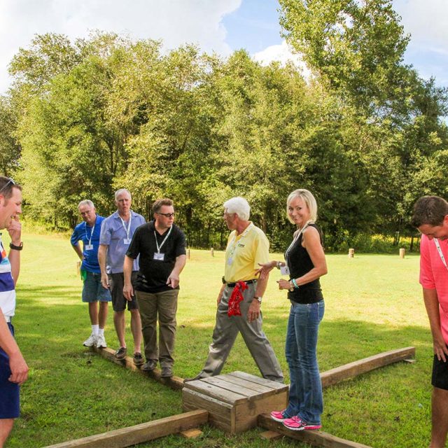 A group of seven adults participates in an outdoor team-building exercise on a wooden platform in a grassy area surrounded by trees during their corporate retreat.