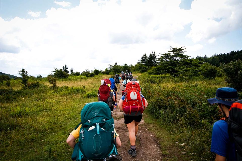 teen campers hike up the hill with full backpacks