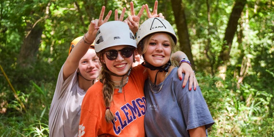 Three smiling individuals pose in a green, wooded outdoor area, each wearing helmets and casual clothing. The person in the middle wears an orange "CLEMSON" shirt. The two people on the ends have their arms around each other, and the person behind gives bunny ears—a perfect snapshot of summer camp camaraderie.