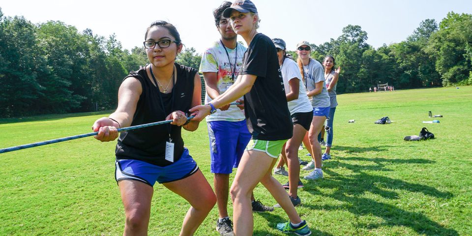 A group of young adults participate in tug-of-war on the sports field at Camp Canaan during a team-building activity.