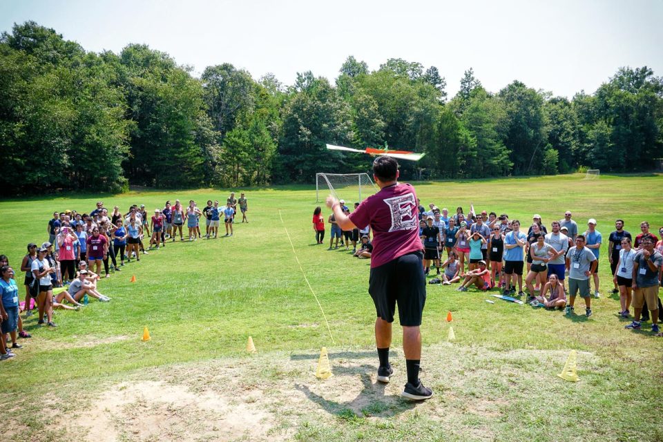 A person wearing a maroon shirt is holding a model airplane, preparing to launch it, with a large group of people gathered around on a grassy sports field.