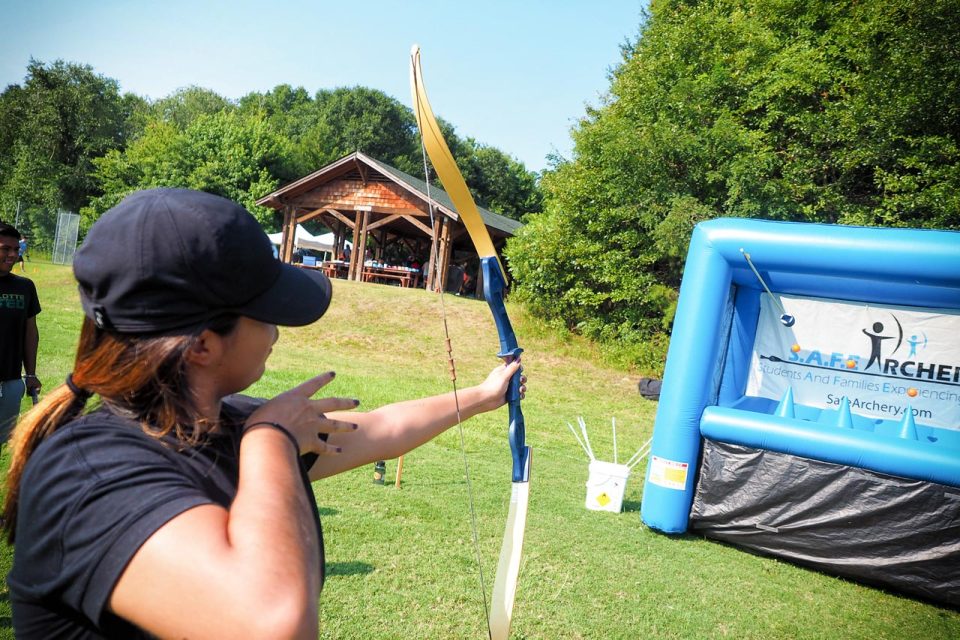 A person wearing a black cap is poised to shoot an arrow from a bow at an archery target. A blue inflatable archery backdrop with "S.A.F.E. Archery" written on it is in the background. A wooden picnic pavilion and green trees are also visible.