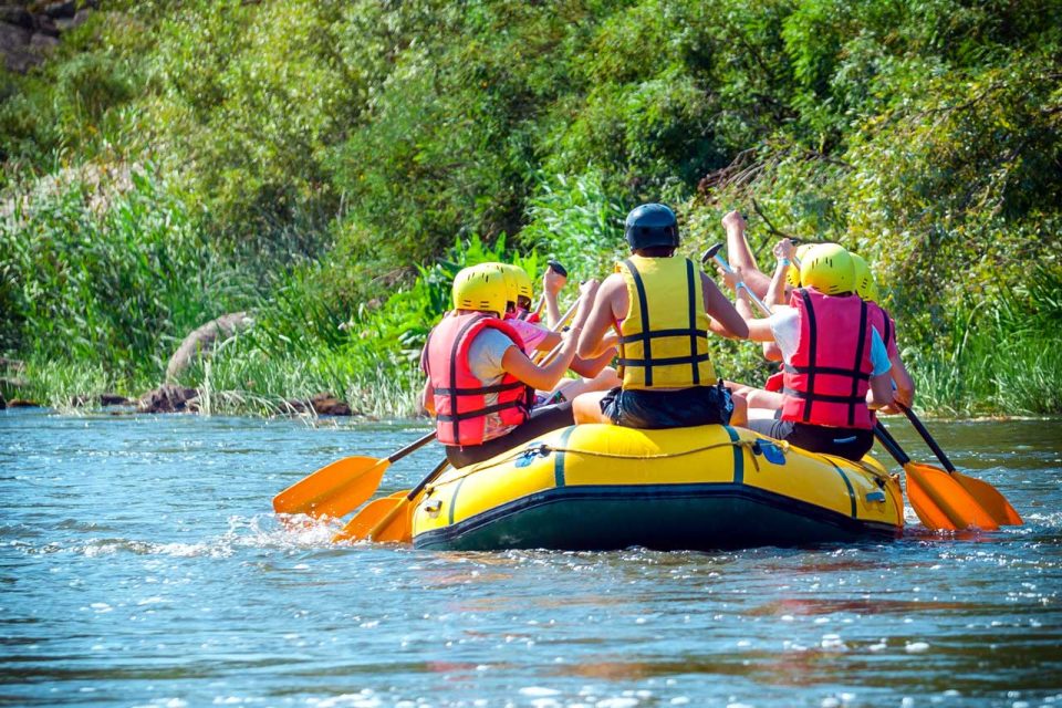 A group of four people wearing helmets and life jackets paddle a yellow inflatable raft down a river surrounded by lush green vegetation.