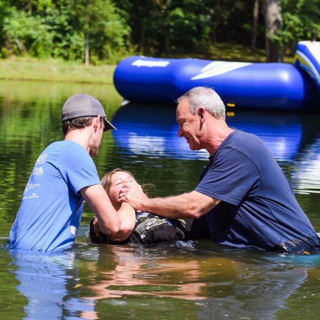 Two men baptize a young girl in the lake at Camp Canaan. The scene takes place in daylight with a large inflatable structure in the water, and trees line the shore.