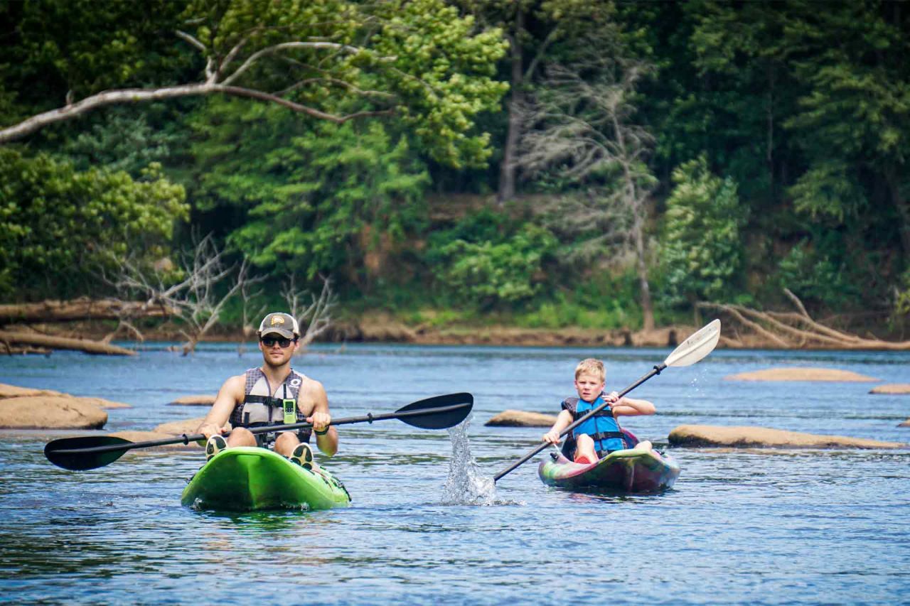 Two people are kayaking on the Catawba river surrounded by greenery, as part of a summer camp activity. The person on the left is an adult with a hat and life vest, and the person on the right is a child wearing a life vest. Both are paddling through the water on a sunny day.