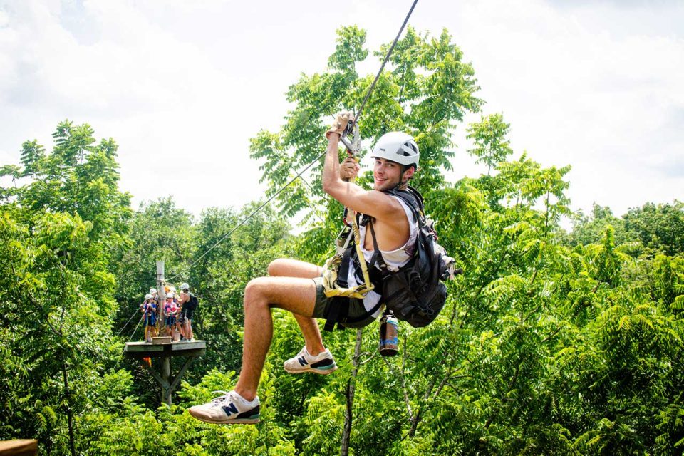 A person wearing a helmet and harness is ziplining through lush, green trees. They are smiling and seem to be enjoying the outdoor adventure. In the background, a group of people stand on a platform, waiting for their turn on the zipline.