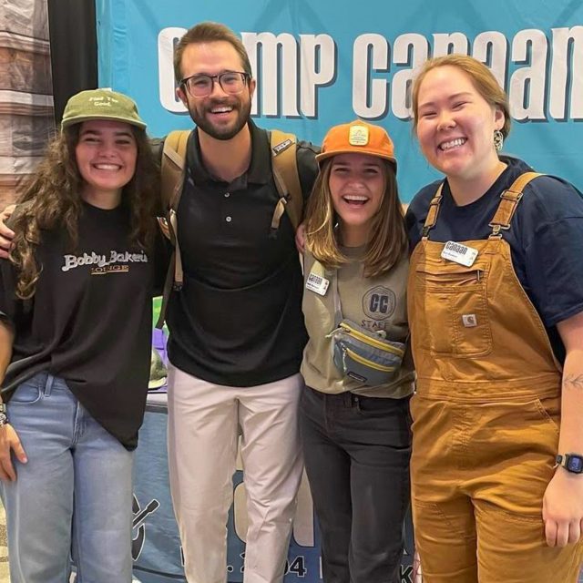 Four smiling people stand in front of a "Camp Canaan" banner at a recruiting event. They are posing together with their arms around each other, embodying the spirit of Camp Canaan.