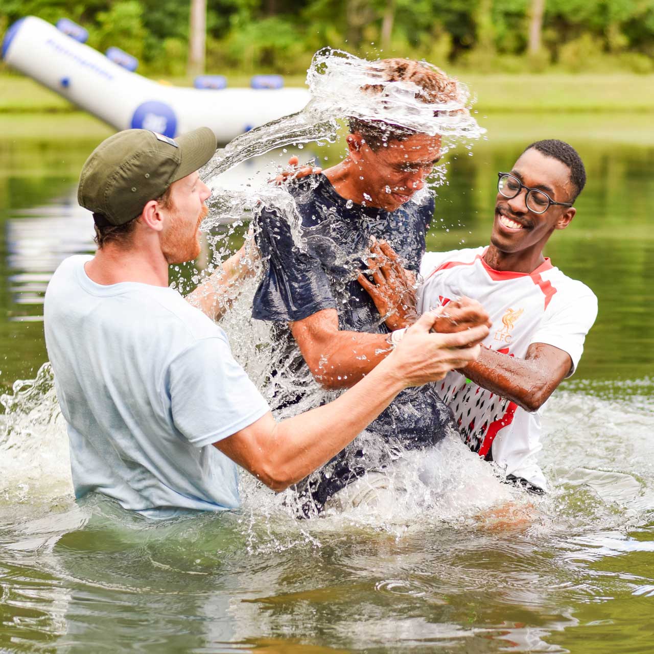 Two male counselors baptize a fellow male counselor in Lake Canaan. The water whips off his hair as he comes up out of the lake. Everyone smiles with excitement.