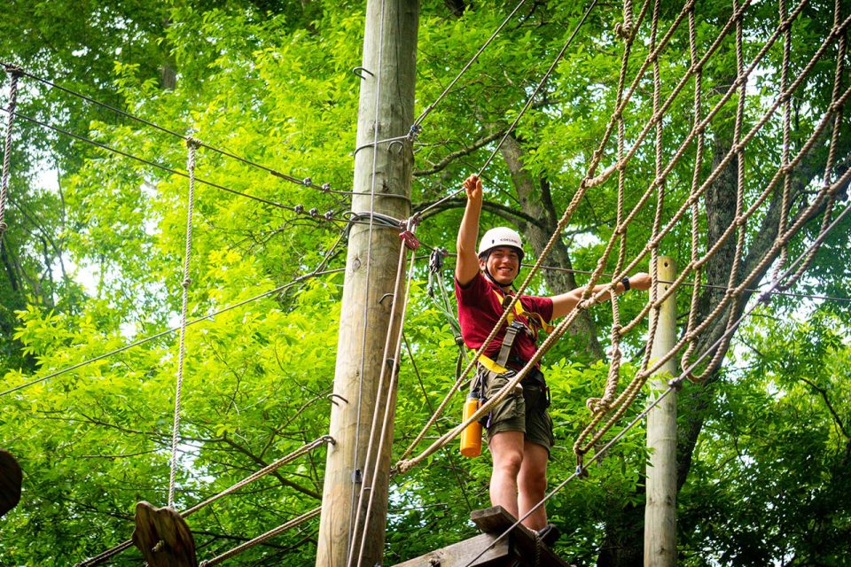 A male adventure guide wearing a helmet and safety harness is climbing a high ropes course amidst lush green trees at Camp Canaan. They are holding onto ropes.