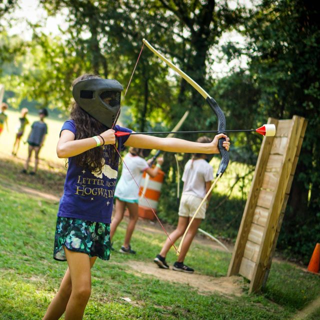 A camper wearing a protective mask and holding an archery tag bow and arrow aims at a target during a summer camp activity.