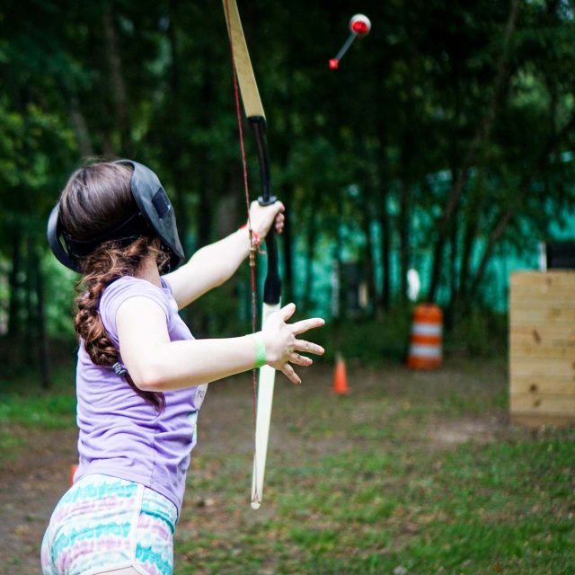 At an overnight summer camp, a camper in a purple shirt and colorful shorts is shooting an arrow from an archery tag bow in an outdoor setting. They are aiming carefully while wearing a protective mask.