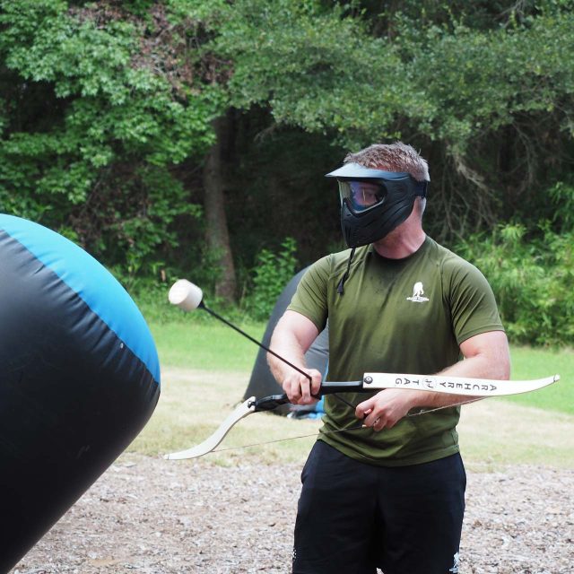 A man stands outdoors at an overnight summer camp, wearing a face mask and an olive-green shirt, holding a bow equipped with a foam-tipped arrow. They are in the middle of a game of archery tag.