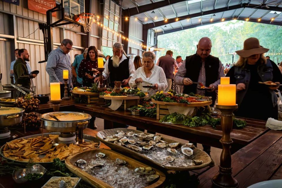 A group of people are gathered around a buffet table in the covered basketball court at Camp Canaan with string lights overhead. The table is laden with various foods, including oysters, dips with crackers, and pretzel bites. People are serving themselves with plates in hand.