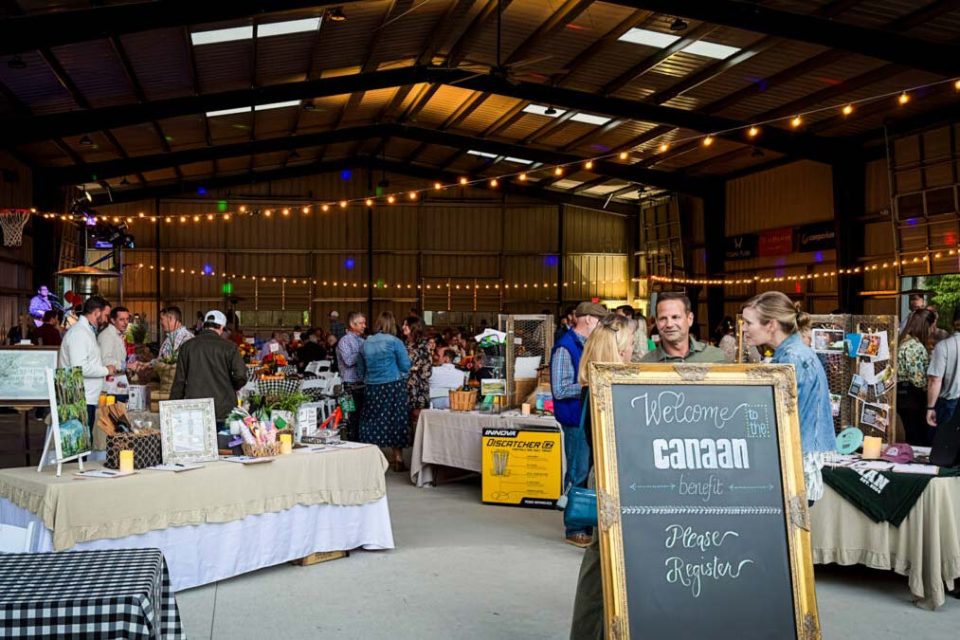 People gather in the covered basketball court at Camp Canaan for the Camp Canaan benefit featuring various silent auction items with handcrafted goods and decorations. Attendees are mingling and browsing items. A chalkboard sign in the foreground reads, "Welcome to the Camp Canaan benefit. Please register." String lights add a warm ambiance.