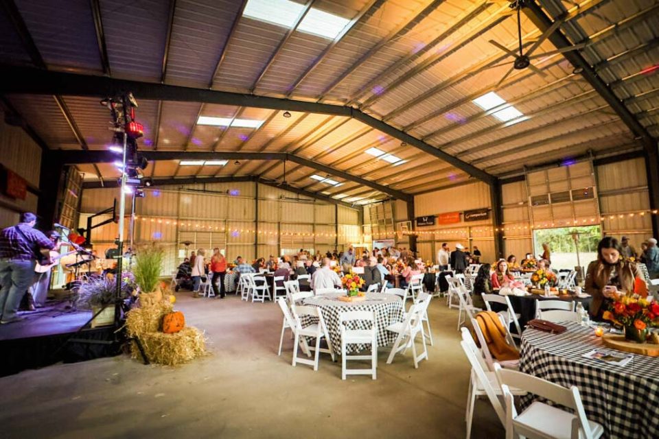 A large indoor event at Camp Canaan features people seated at tables covered in checkered tablecloths. Decorations include pumpkins and autumn-themed centerpieces. A stage on the left hosts a live band performing. The venue is brightly lit with string lights and colorful spotlights.