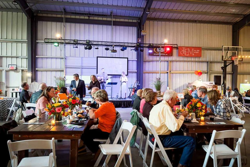 People are seated at wooden tables decorated with bouquets of flowers, attending an event in the covered basketball court at Camp Canaan. A stage at the front has speakers and a screen. String lights are hung for ambiance. Conversations and dining are taking place.