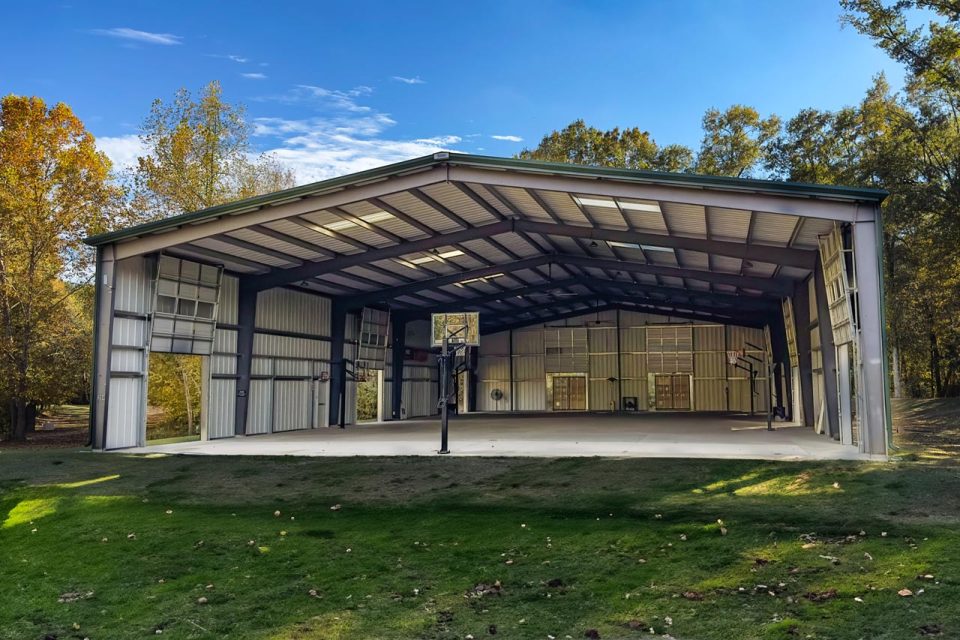 At Camp Canaan, an open-air metal structure with a pitched roof and large doorway openings on each wall houses a basketball court with multiple hoops. The building is surrounded by green grass and trees, under a clear blue sky, providing the perfect setting for an overnight summer camp and retreat center.