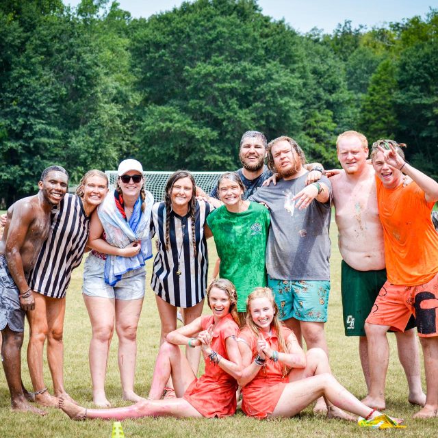 A group of summer camp counselors, some covered in mud, stand and kneel on grass at Camp Canaan, with trees in the background. They smile and pose for the camera, with two sitting down in playful poses. It appears to be a fun, messy outdoor event during summer camp.