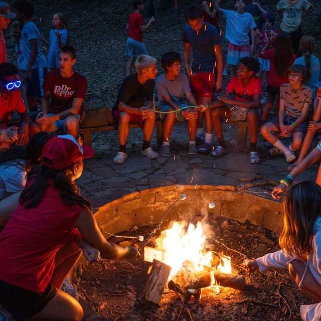 A group of people, mostly children and teenagers, are gathered around a campfire at Camp Canaan at night. They are engaged in conversations and roasting marshmallows. The scene is lively and the fire illuminates their smiling faces.