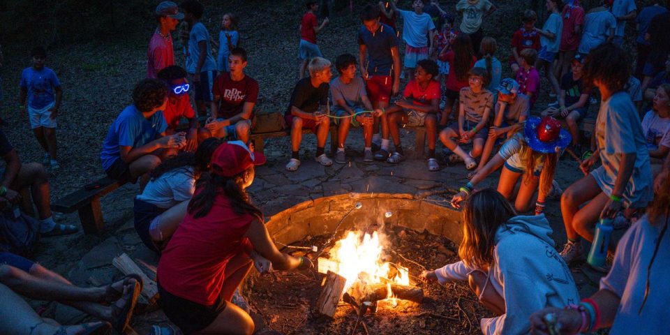 A group of people, mostly children and teenagers, are gathered around a campfire at Camp Canaan at night. They are engaged in conversations and roasting marshmallows. The scene is lively and the fire illuminates their smiling faces.