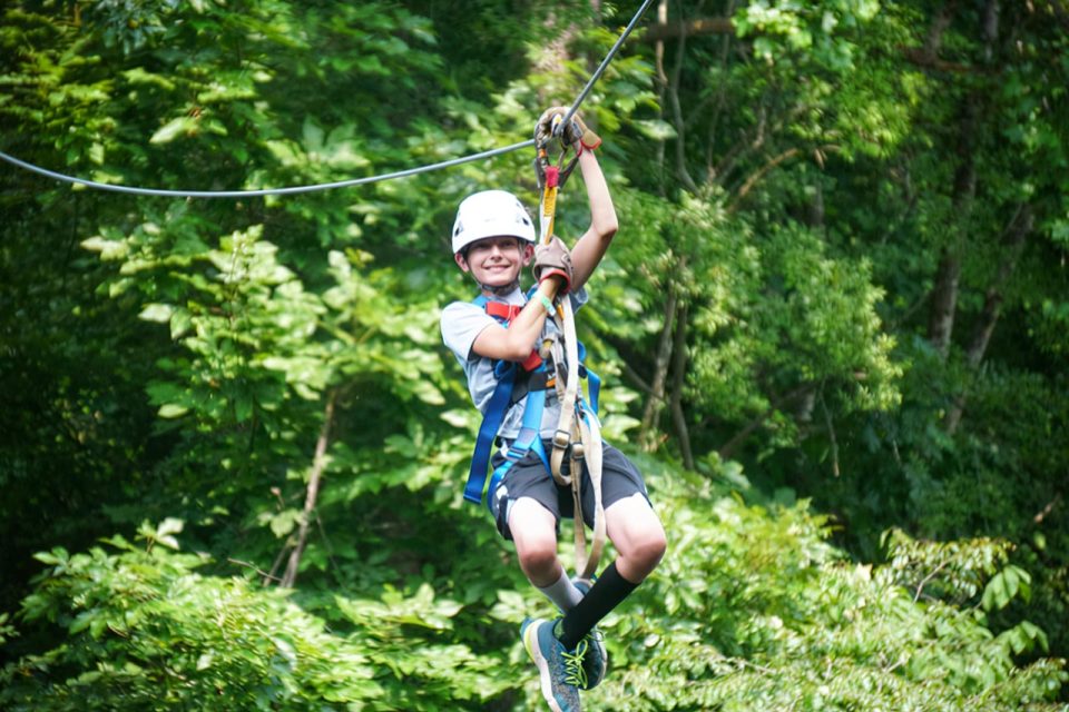 A child wearing a helmet and harness is ziplining through lush, green trees at Camp Canaan. They are smiling and holding onto the zipline equipment as they glide above the dense trees below.