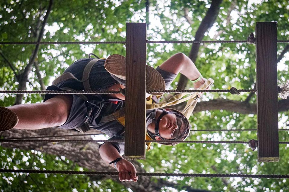 A person wearing a helmet and safety harness is seen from below navigating a high ropes course element in a wooded area at Camp Canaan. They are holding onto ropes while navigating wooden planks. The background is filled with green leaves from the trees overhead.