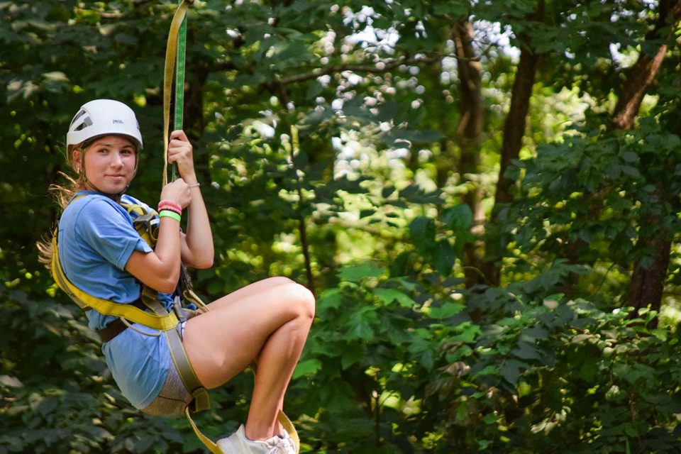 A young person wearing a helmet and harness rides a zipline through a wooded area, surrounded by lush green trees and foliage. They are dressed casually in a blue shirt and shorts, appearing to enjoy the zipline during summer camp.