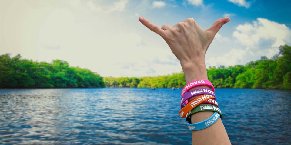 A hand with multiple colorful wristbands is making a shaka sign in the foreground of a serene river surrounded by trees under a partly cloudy sky. The wristbands say Canaan Hover.