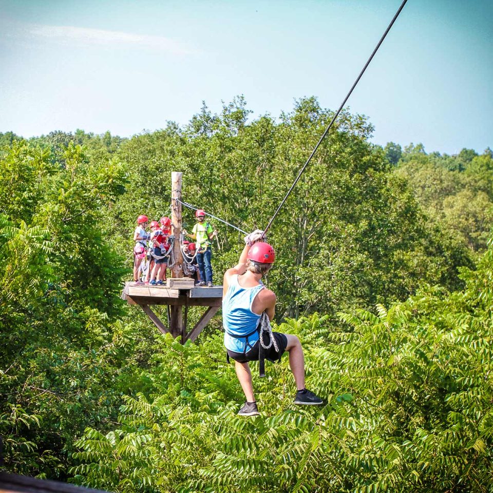 A person wearing a blue tank top and red helmet rides a zipline through the lush green trees Camp Canaan.