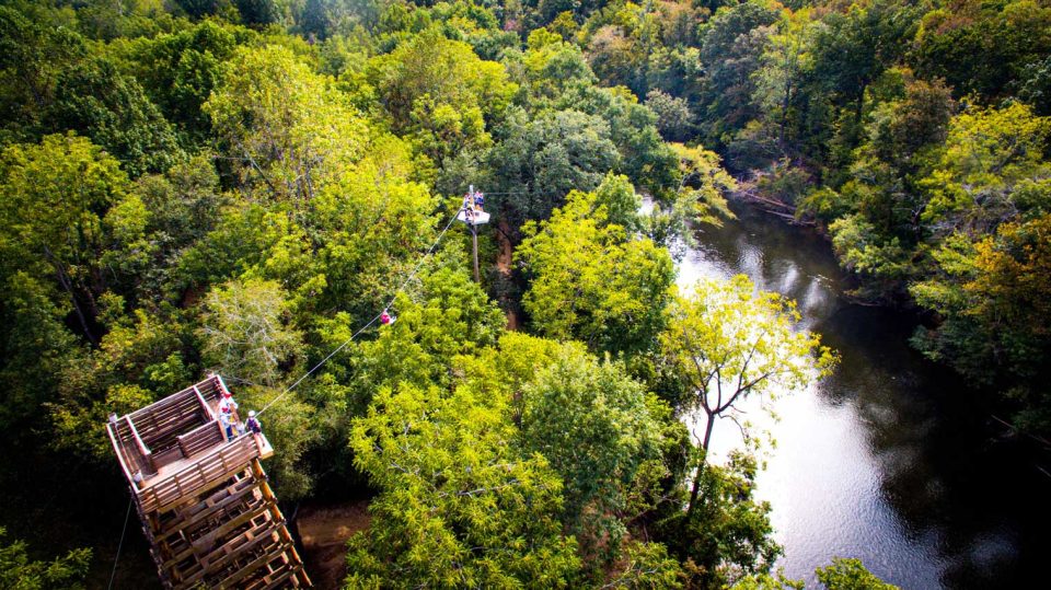 Aerial view of a lush, green forest with the Catawba river winding through it. The Zipline Canopy tour tower stands on the left at where people are ziplining across the river to a platform on the other side.