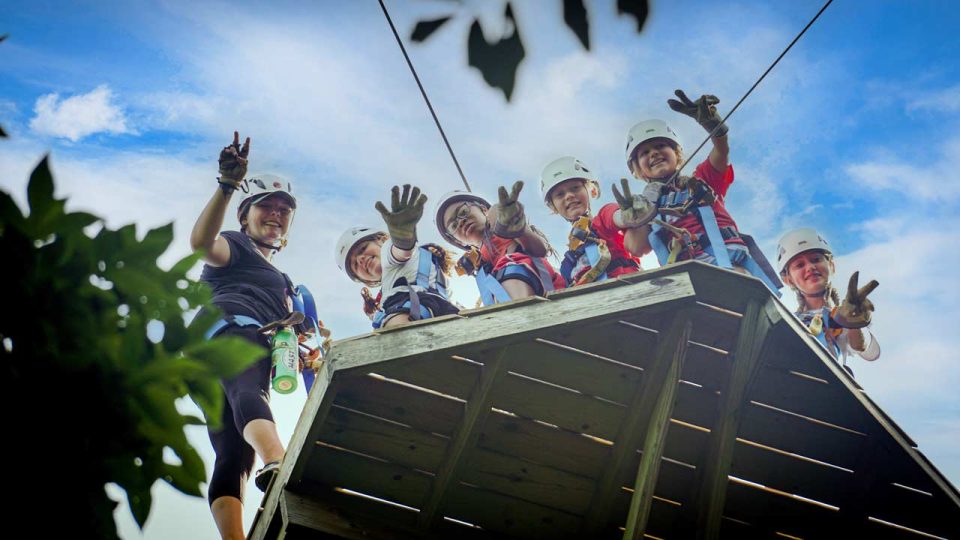 A group of six people wearing helmets and safety gear stand on a Zipline Canopy Tour platform at Camp Canaan, smiling and giving peace signs. The sky is bright blue with some clouds.