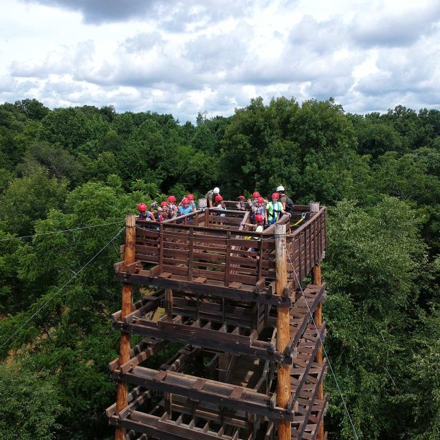 A group of people wearing helmets and harnesses stand on the Zipline Canopy Tour tower in the middle of a wooded area at Camp Canaan, high above the ground.