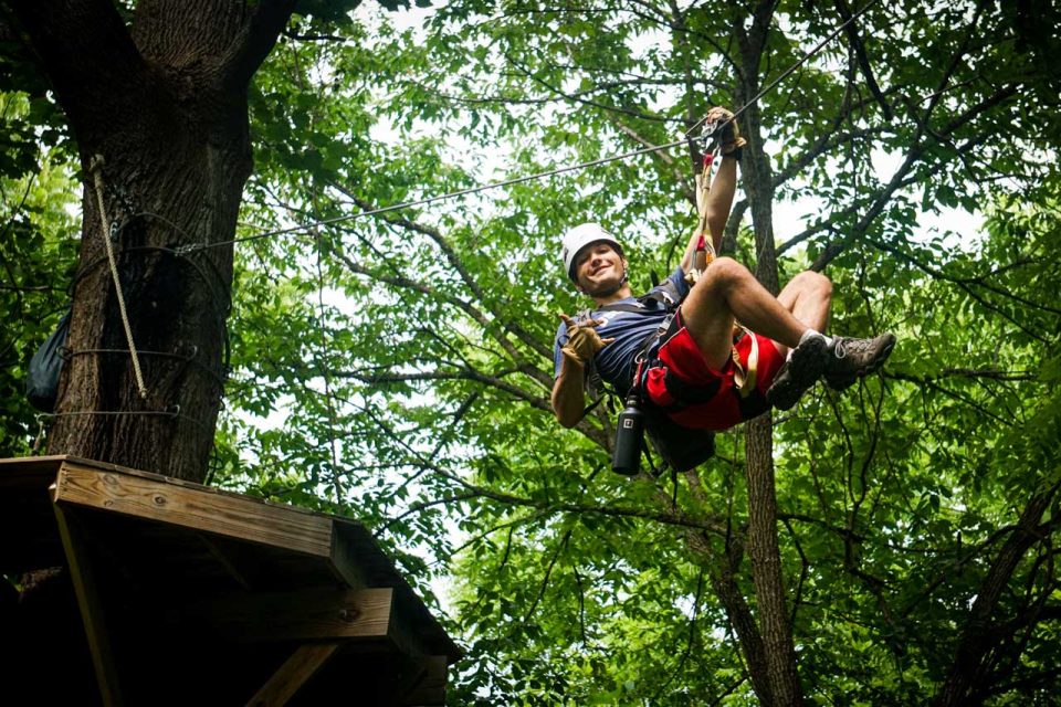 An adventure guide at Camp Canaan is ziplining through a wooded area, wearing a helmet and safety harness. They are smiling and giving a thumbs-up, while surrounded by lush green foliage and trees.