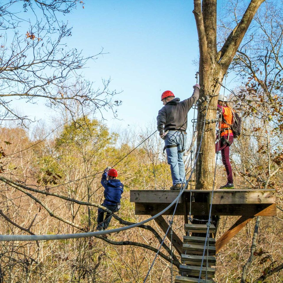 Three individuals stand on a wooden Zipline Canopy Tour platform high in a tree at Camp Canaan. Clad in helmets and harnesses, they enjoy the thrill surrounded by leafless trees on a clear day. One person zips across while two others prepare on the platform.