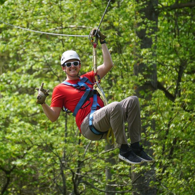 A person wearing a red shirt, brown pants, gloves, white helmet, and sunglasses is ziplining through a forest canopy at Camp Canaan. They are smiling, raising one arm with a thumbs-up gesture while secured with harnesses and safety equipment. Green foliage is visible behind them.